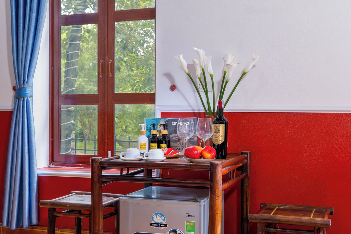 A room with a window, a table with a vase of flowers, a refrigerator, and a red wall.