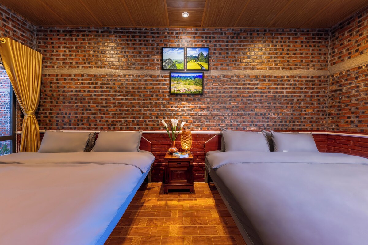 A hotel room with a brick wall, two beds, a nightstand with a lamp, and a window with curtains.