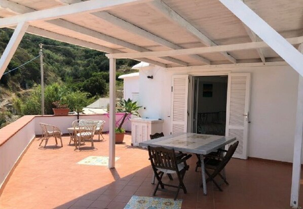 A covered outdoor patio with a table and chairs, a potted plant, and a view of greenery in the background.