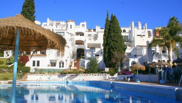 A swimming pool with a thatched umbrella, white buildings with balconies, and palm trees in the background.