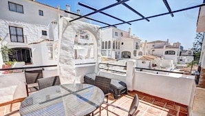 A rooftop terrace with a round glass table and wicker chairs, white buildings in the background, and a clear blue sky.