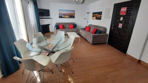 A modern living room with a glass dining table, white chairs, a grey sofa with red and blue cushions, a flat-screen TV, and a fireplace.