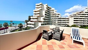 Eine Dachterrasse mit Gartenmöbeln, Blick auf das Meer und ein modernes Gebäude im Hintergrund.