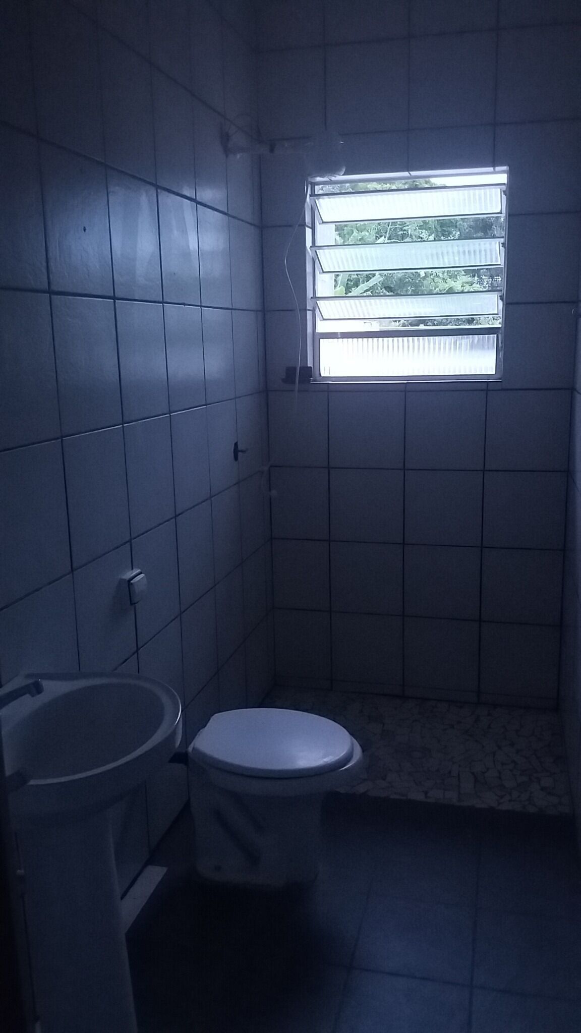 A small, dimly lit bathroom with a toilet, sink, and a window with blinds.