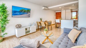 A modern living room with a grey sofa, a dining area with a wooden table and chairs, a kitchen with wooden cabinets, and a large window. - Luxury apartment Blue Sea in Alcudia beachview (Alcúdia)