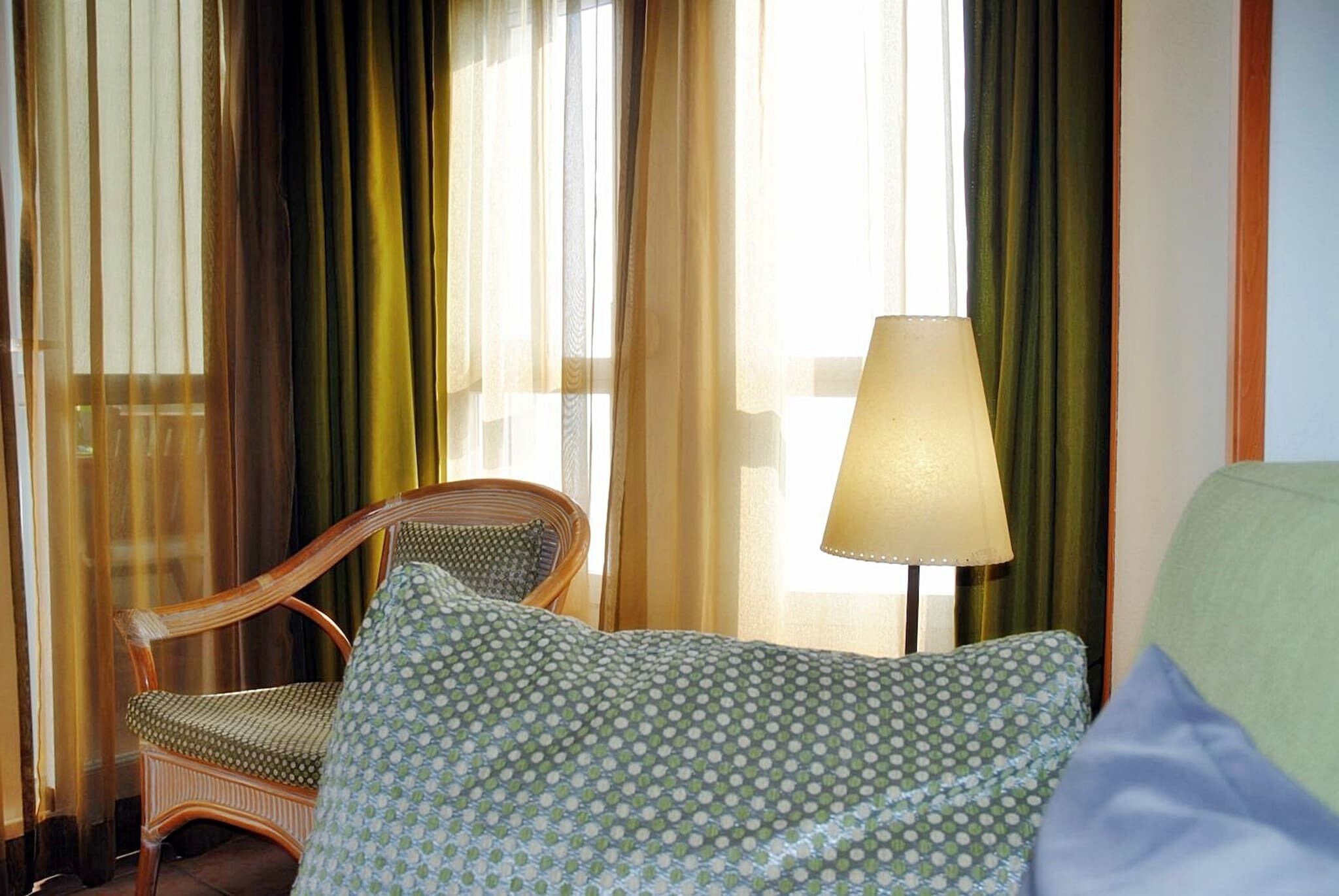 A hotel room with a bed, a chair, a lamp, and sheer curtains.