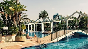 Ein Swimmingpool mit einer Brücke, umgeben von Palmen und einem überdachten Bereich mit weißen Säulen.