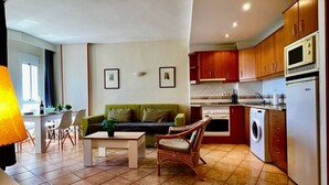 Eine moderne Küche mit Holzschränken, einem eingebauten Backofen, einer Mikrowelle und einer Waschmaschine. Es gibt einen Essbereich mit Tisch und Stühlen sowie ein Wohnzimmer mit einer Couch und einem Korbstuhl.