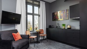 A modern living room with a dark sofa, a coffee table, and a TV. There's a window with curtains, a painting on the wall, and a kitchen area with a sink and cabinets. - Hotel Filip (Oslo)
