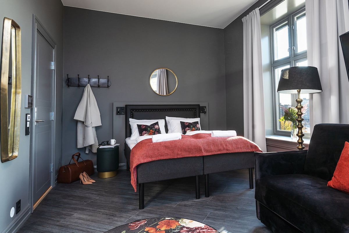 A modern hotel room with a large bed, a black armchair, a flat-screen TV, and a window with curtains.