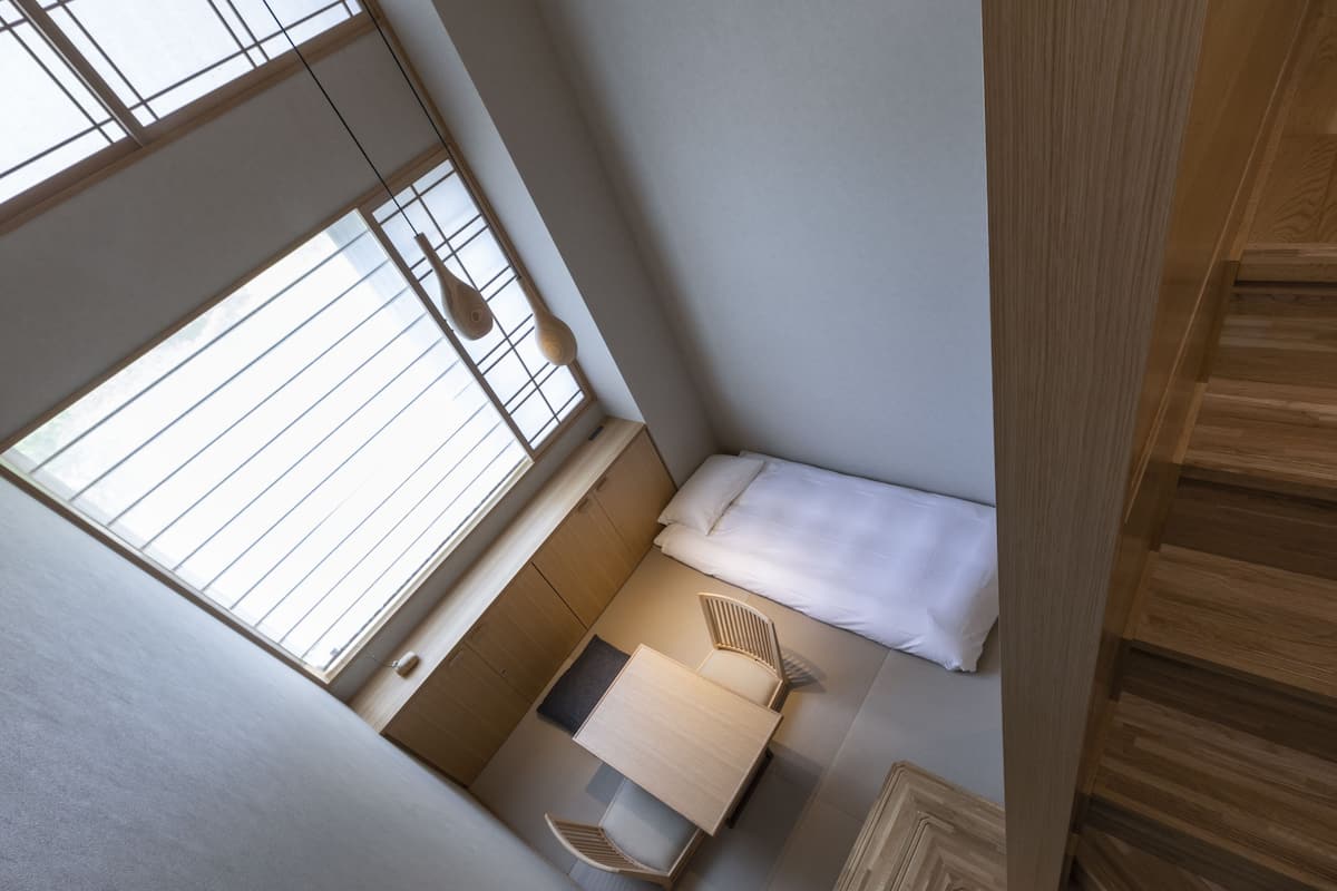 A compact bedroom with a bed, a small table, and a chair, featuring a large window and wooden flooring.