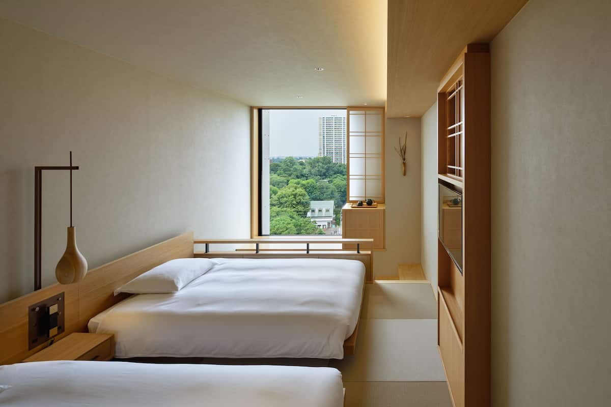 A modern hotel room with two beds, a large window offering a city view, and a minimalist design.