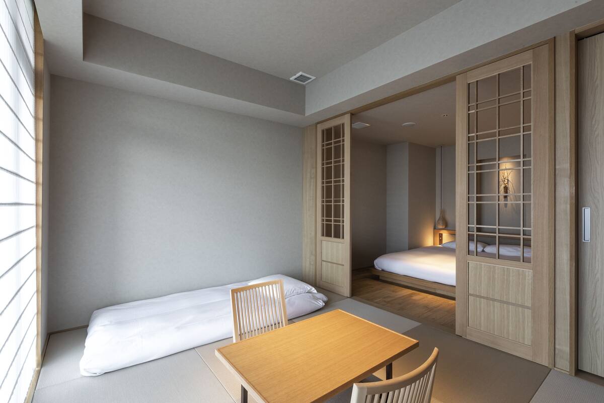 A compact room with a bed, a table, and a chair. There is a sliding door leading to another room with a bed.
