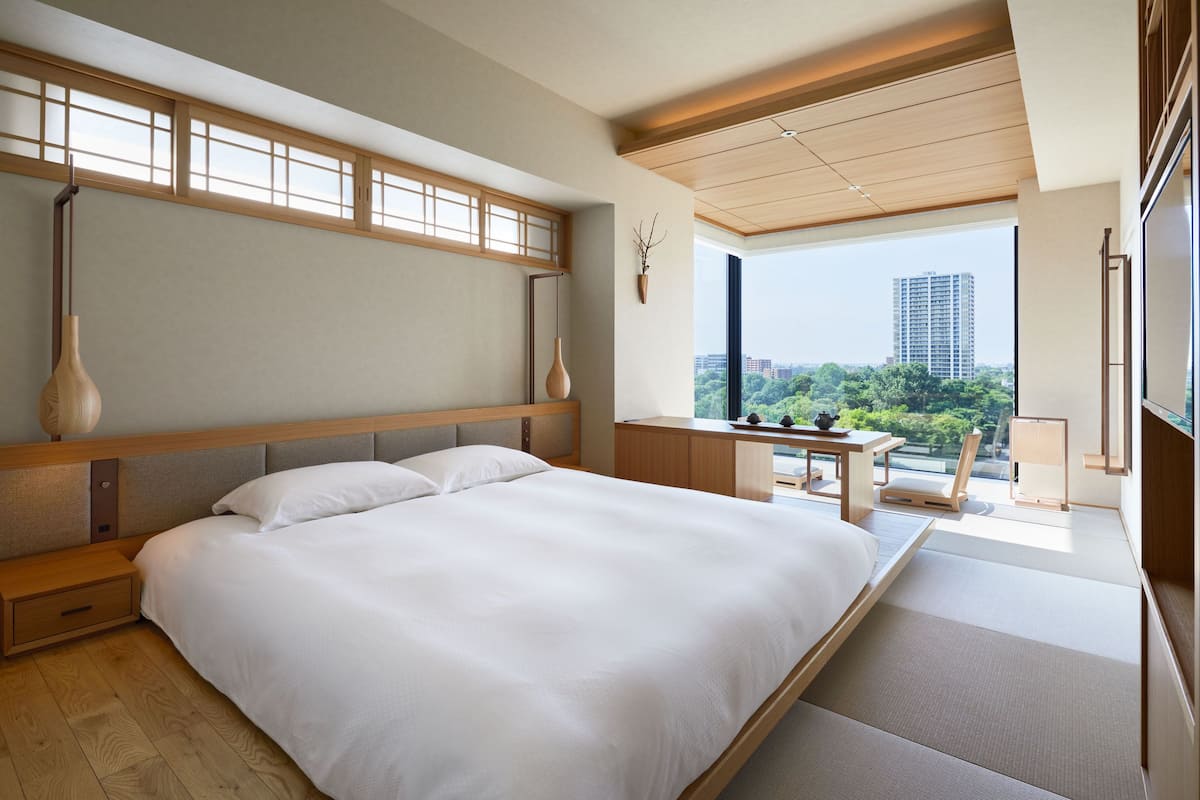 A modern hotel room with a large bed, a wooden headboard, a small desk, and a view of a cityscape through large windows.