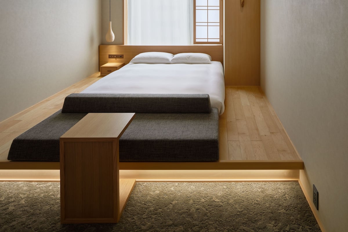 A modern bedroom with a large bed, a bedside table, a nightstand, a floor lamp, and a window with sheer curtains.