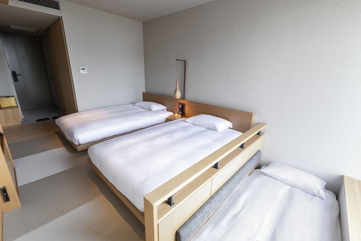 A hotel room with two single beds, a wooden headboard, a bedside table with a lamp, and a door leading to a hallway.