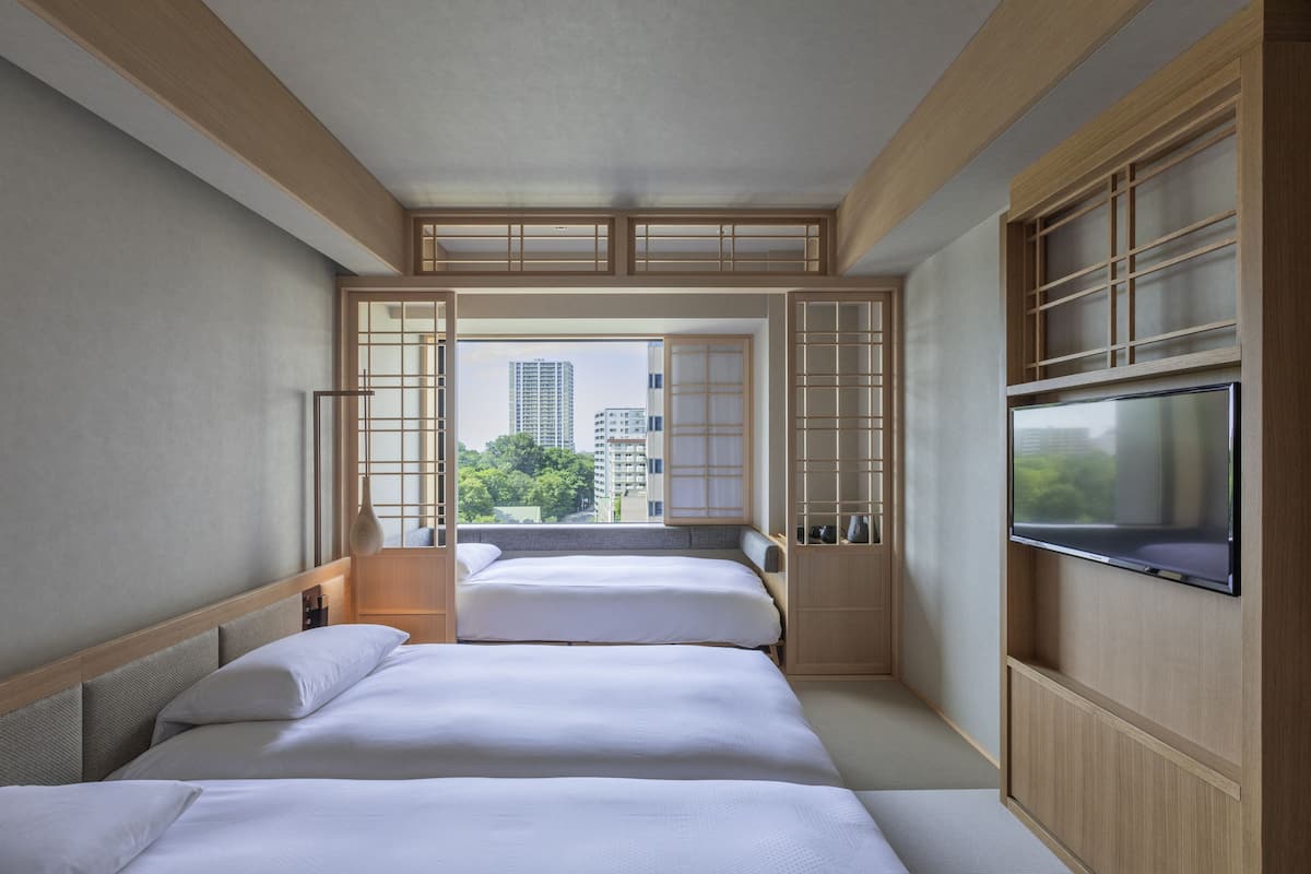 A modern hotel room with two beds, a TV, and a view of the cityscape.