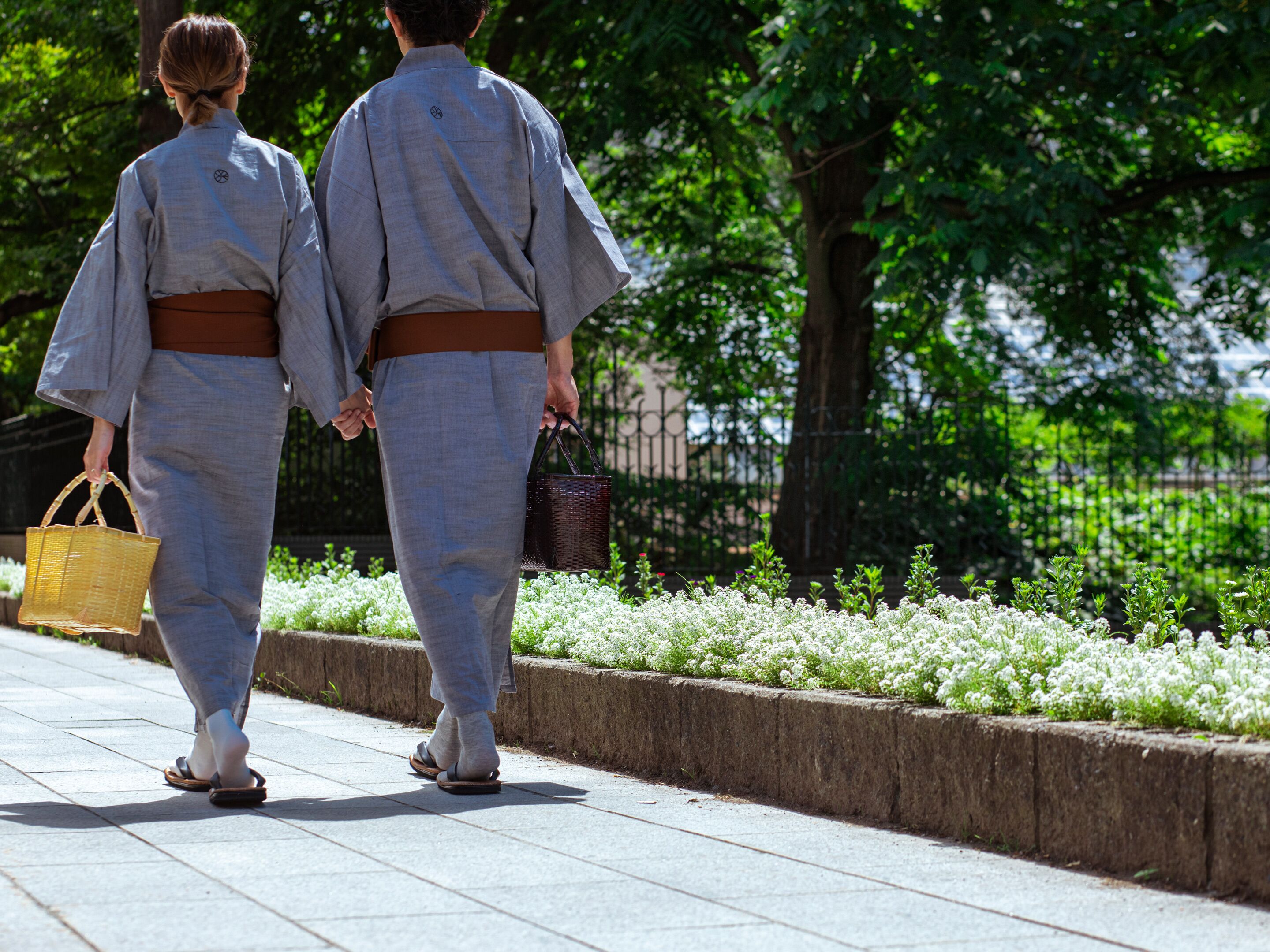 Two individuals in traditional attire walking on a pathway with a garden and a fence in the background.