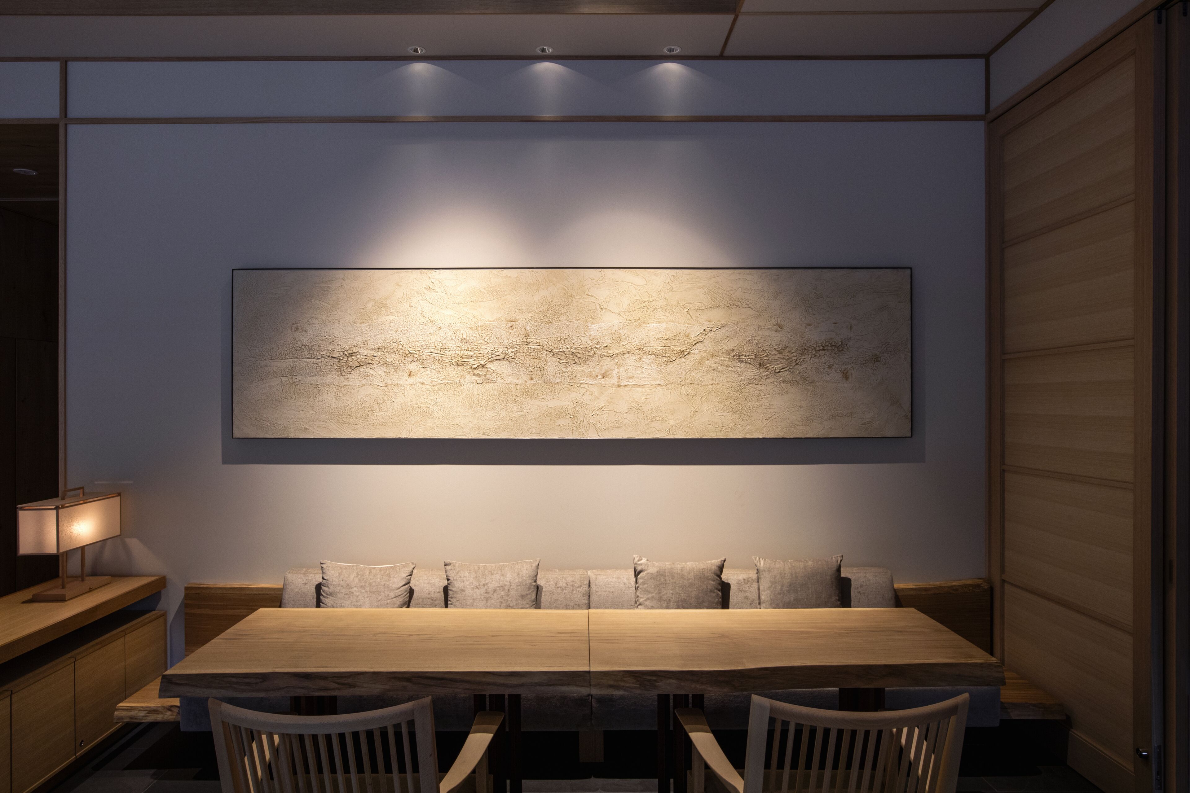 A dining area with a long wooden table, cushioned benches, and a large abstract painting on the wall.