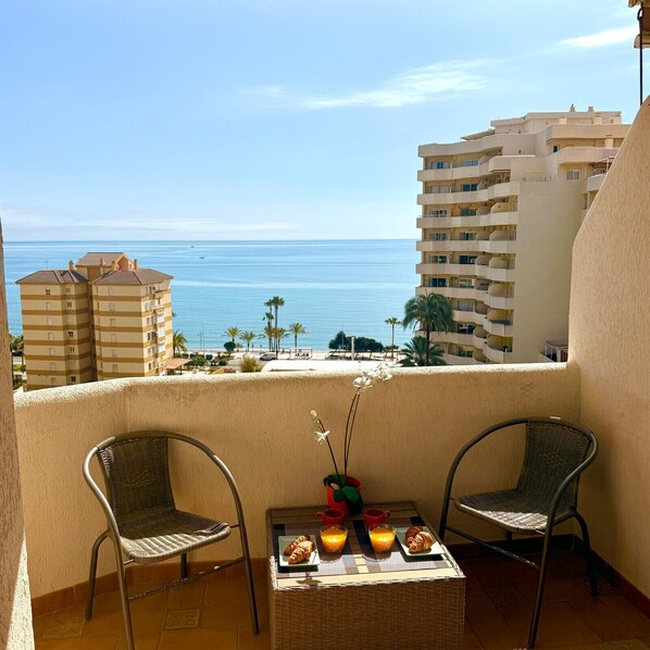 A balcony with two wicker chairs, a small table with croissants and orange juice, and a view of the ocean and buildings. - Spacious Studio in Benalbeach with sea views (Benalmádena)
