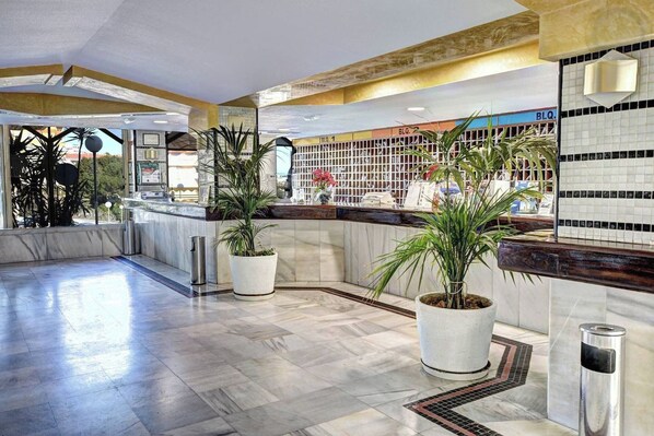 A lobby with marble flooring, potted plants, a reception desk, and a tiled wall with a grid pattern.