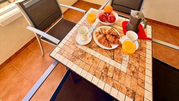 Een balkon met een ontbijttafel, compleet met croissants, fruit en koffie, met uitzicht op de kust.