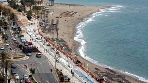 Una carretera costera con una playa a un lado, un camino con coches y un velero en el agua.