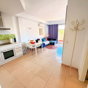 Eine moderne Küche mit integriertem Backofen, ein Essbereich mit weißem Tisch und Stühlen, eine Couch mit bunten Kissen und ein Blick durch eine Glastür auf einen Balkon.