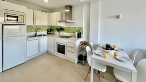 Una cocina moderna con armarios blancos, horno empotrado, microondas y un fregadero. Hay un comedor con una mesa preparada para dos personas, y una planta en el alféizar de la ventana.