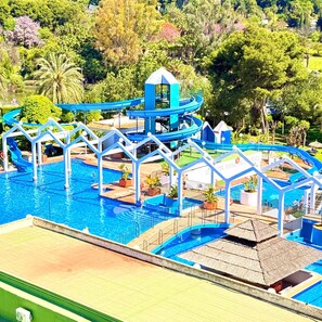Una piscina con tobogán de agua, rodeada de follaje y una estructura con techo de paja.