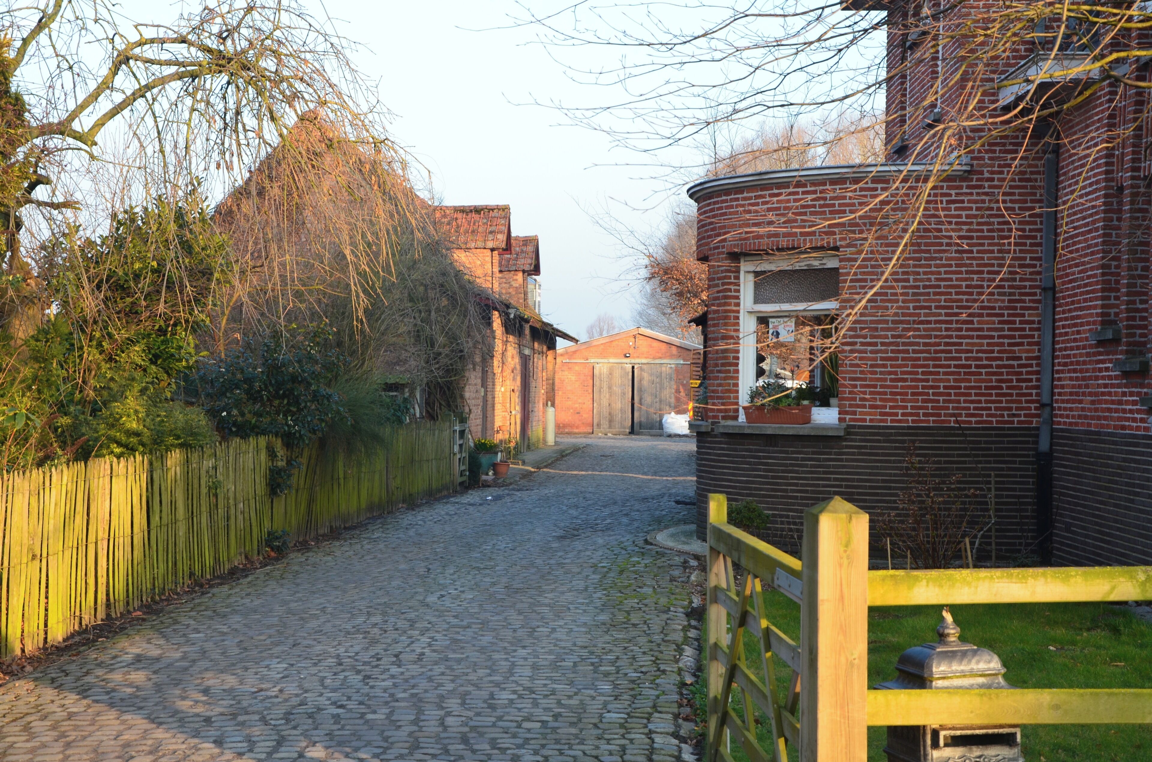 Une ruelle pavée bordée d’immeubles en briques, avec une clôture en bois et un portail jaune.