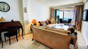 A hotel room with a sofa, a dining table, a TV, and a view of the sea.