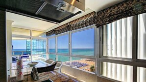 A room with a large window overlooking a beach, a dining area with a table and chairs, and a view of the ocean.
