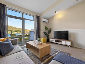 En moderne stue med sofa, sofabord, fjernsyn og udsigt til sø og bakker.