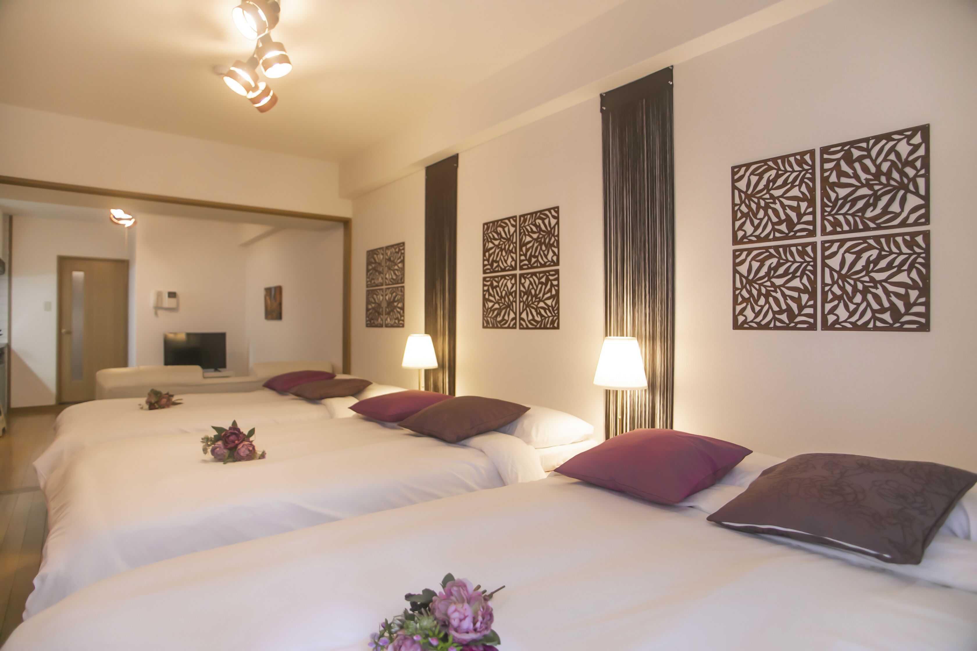 A hotel room with three beds, each with white linens and decorative pillows. There are wall-mounted lamps and a flat-screen TV on a stand.