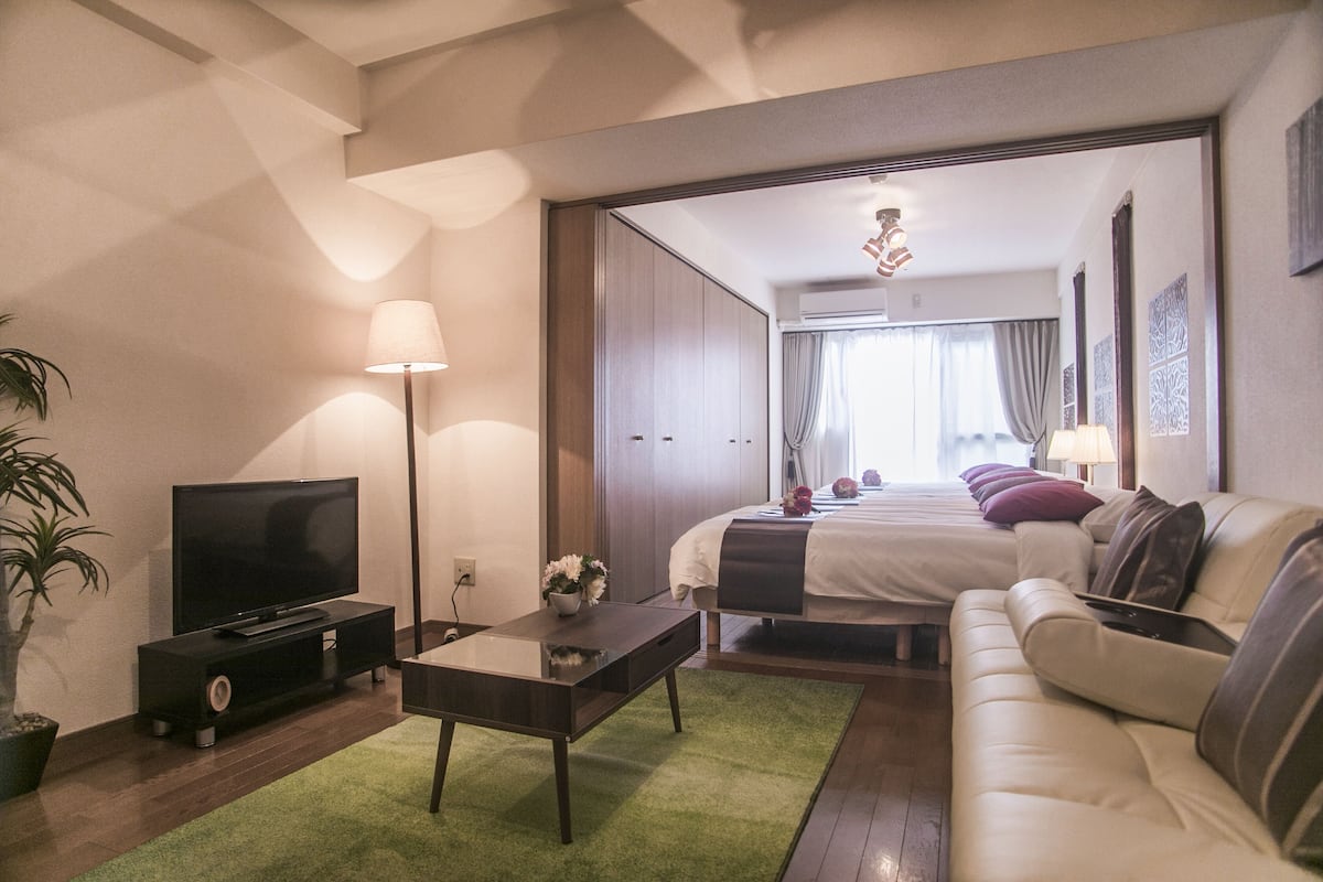 Quarto de hotel moderno com TV de tela plana, sofá, cama com várias almofadas, mesa de cabeceira e tapete verde.