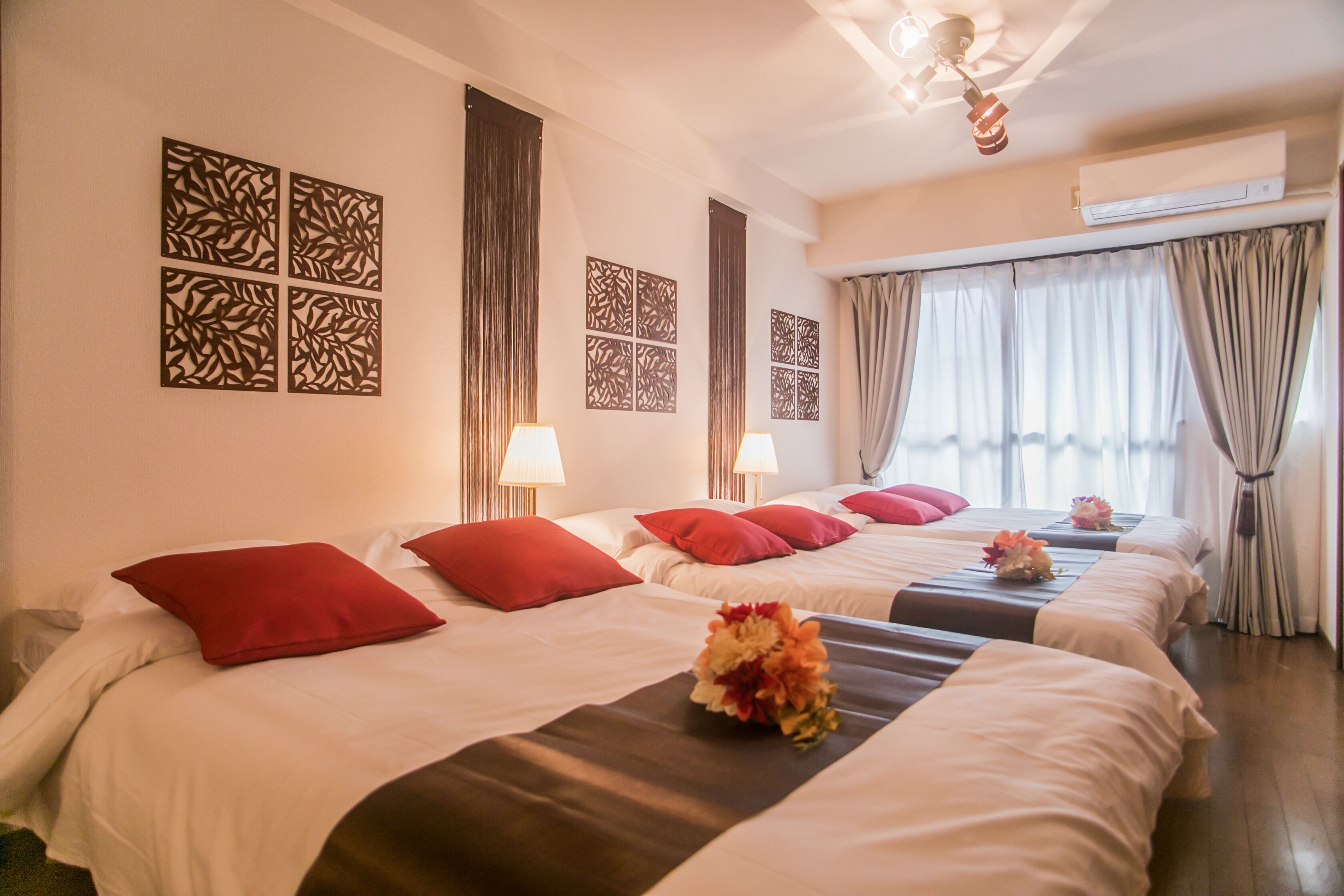 A hotel room with three beds, red pillows, decorative wall panels, and a ceiling fan.