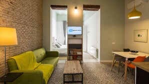 A modern living room with a green sofa, a small wooden table, and a dining area with a white table and orange chairs. A television is mounted on the wall, and there is a brick wall on one side. - Rodrigo Triana 1. 1 bedroom, shared terrace (Sevilla)