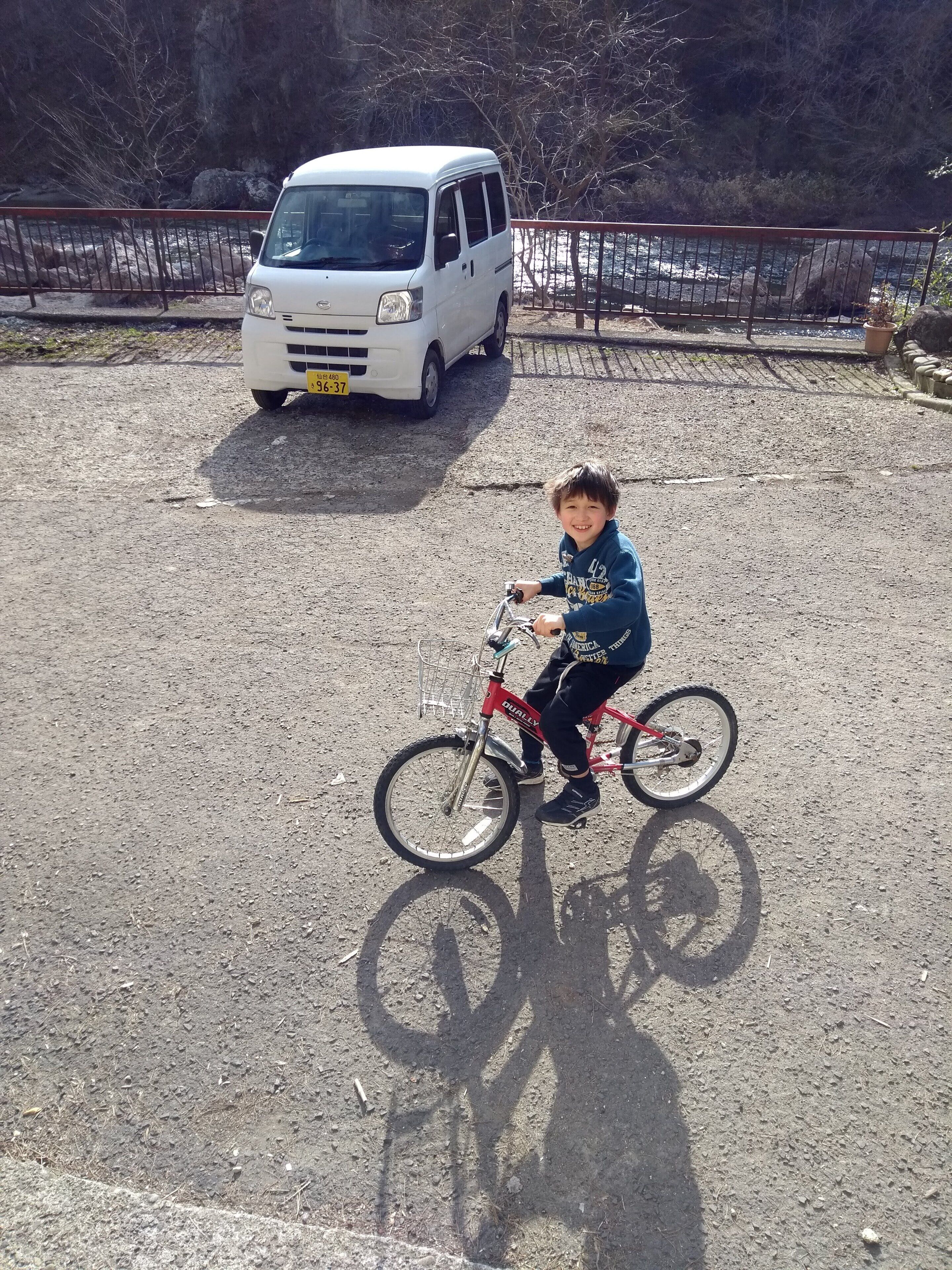 A child riding a bicycle on a gravel path with a white van parked in the background.
