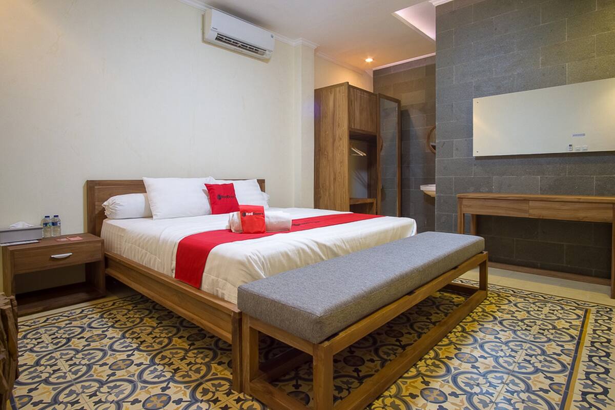 Quarto de hotel com cama, um banquinho, mesa de cabeceira, guarda-roupa e uma pequena mesa.