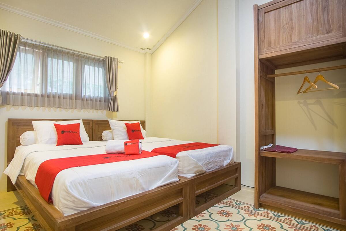 Quarto de hotel com estrutura de cama de madeira, duas camas com roupa de cama branca e detalhes vermelhos, guarda-roupa de madeira e uma janela com cortinas.