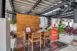 Un espacio interior moderno con una mesa y sillas de madera, plantas en macetas y un cartel informativo rojo.