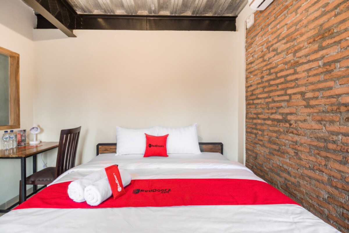 A hotel room with a brick wall, a bed with white bedding and a red pillow, a wooden chair, and a small table with a lamp and water bottles.