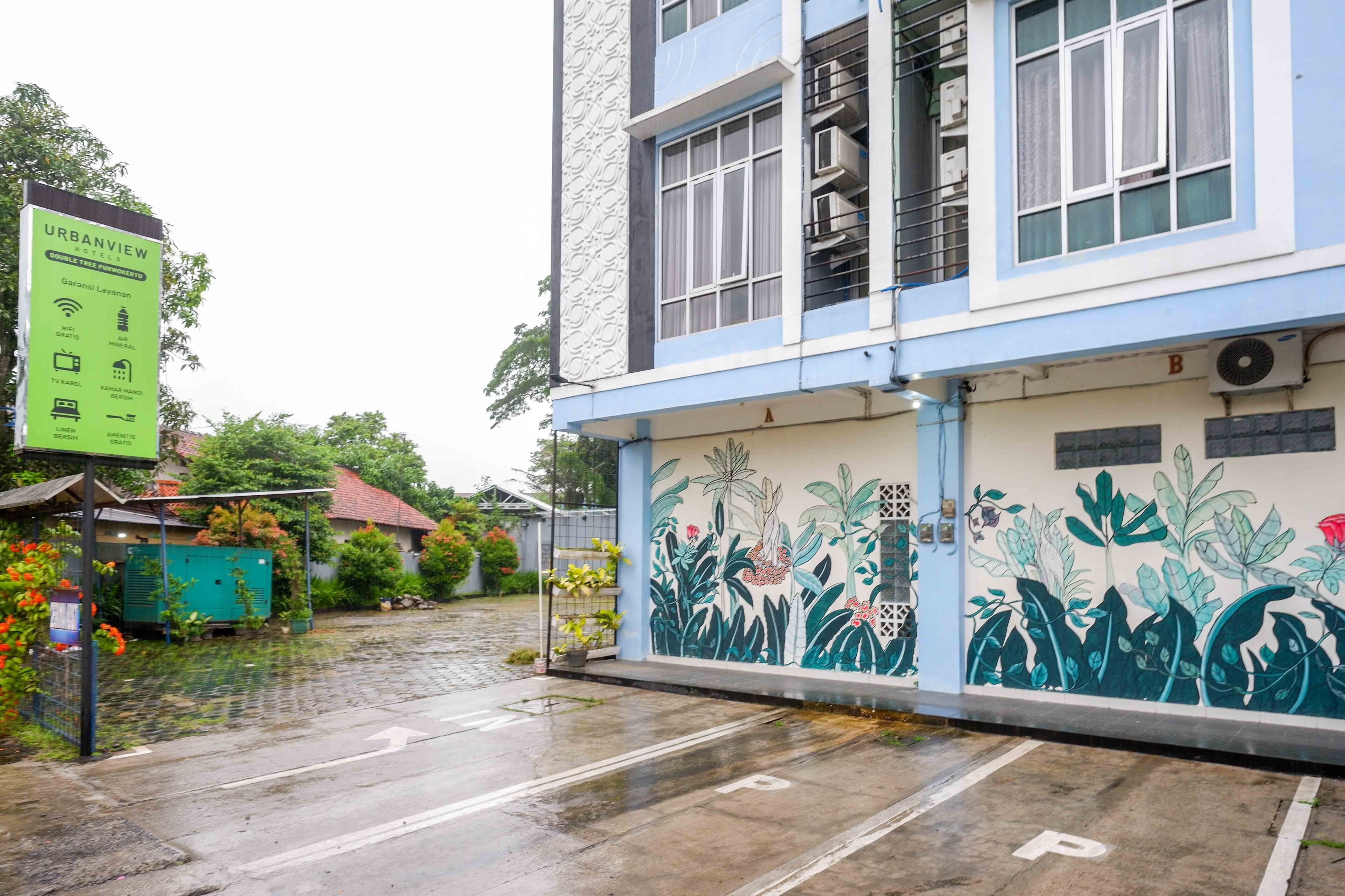 A building with a mural of tropical plants on the side, a green signboard, and a wet street.