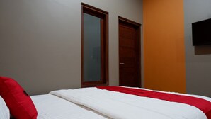 Quarto com cama, travesseiro vermelho, edredom branco, porta de madeira e televisão fixada na parede.