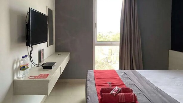 Una habitación de hotel con una cama, un televisor montado en la pared, una mesita de noche con una botella de agua y un objeto rojo, y una ventana con cortinas.