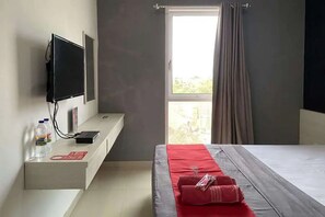 Et hotelværelse med en seng, et fjernsyn ophængt på væggen, et natbord med en vandflaske og en rød genstand, samt et vindue med gardiner.