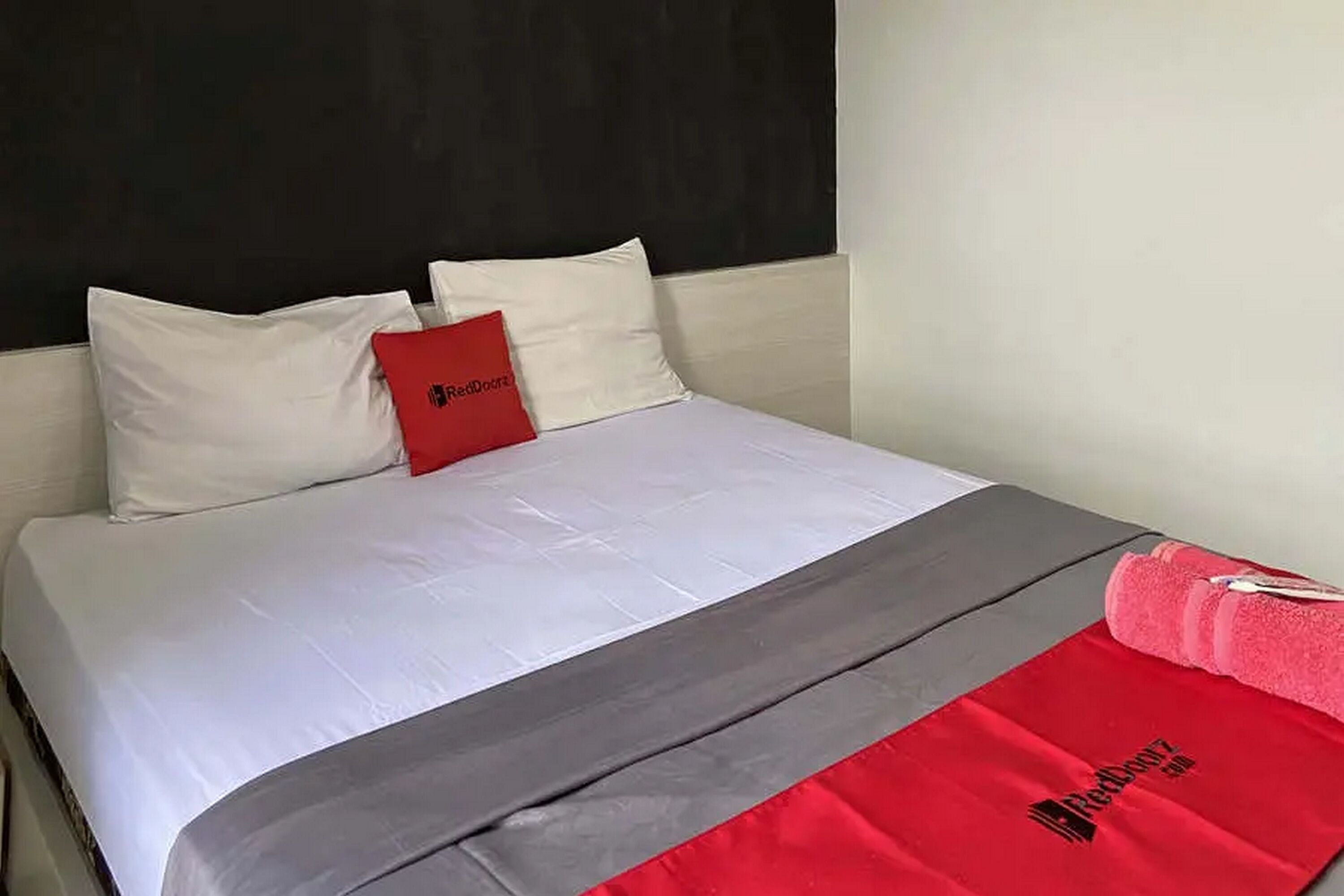Una cama con ropa blanca, una manta gris y roja, y una almohada roja con un logotipo.
