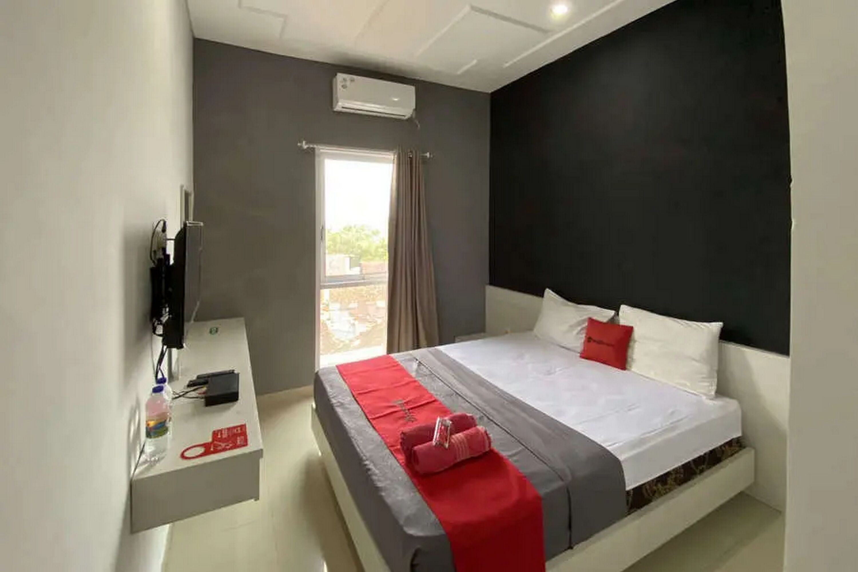 Habitación de hotel con cama, escritorio con teléfono y una botella, ventana con cortinas y unidad de aire acondicionado.