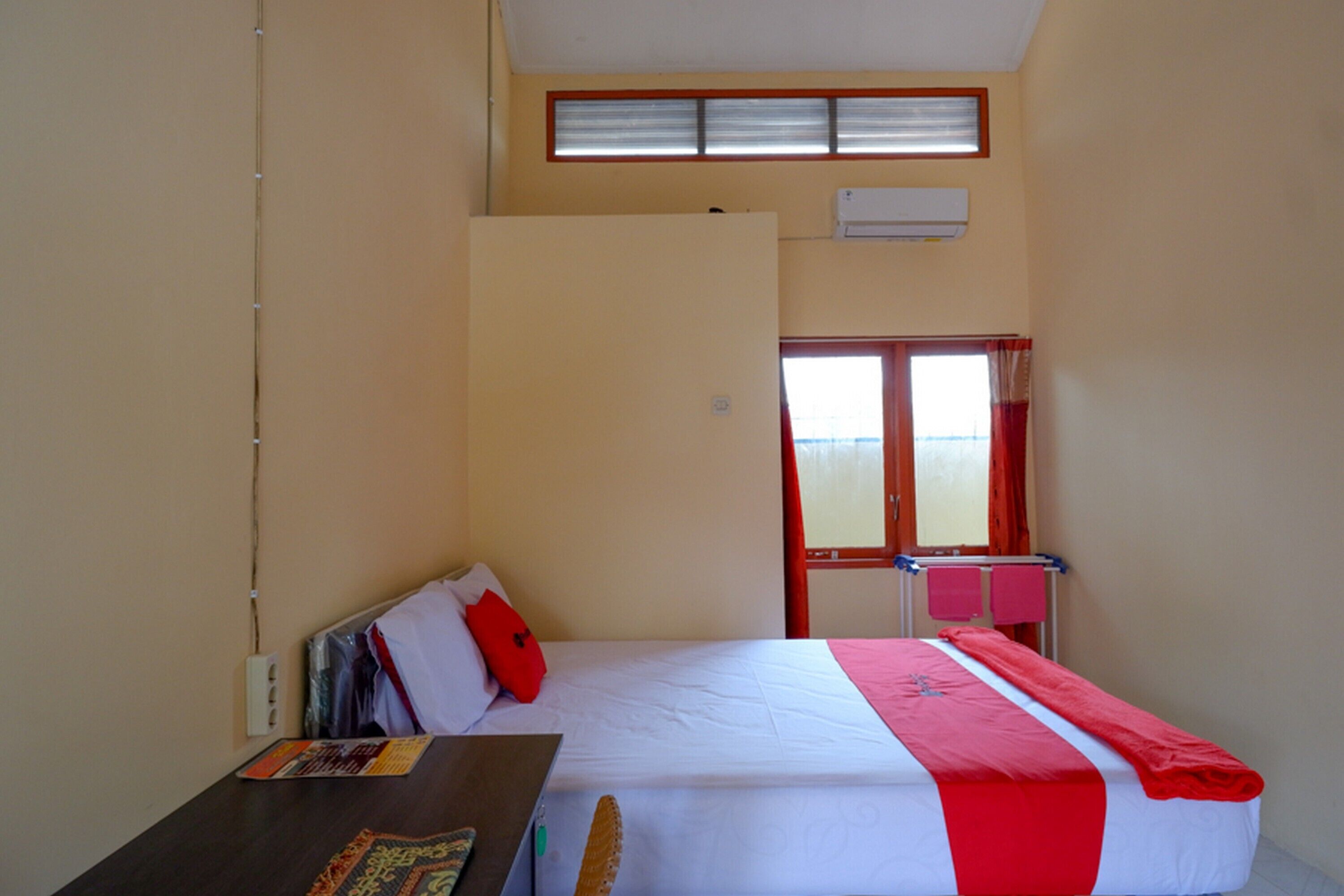 A single-bed room with a window, a desk, and a red and white bedspread.